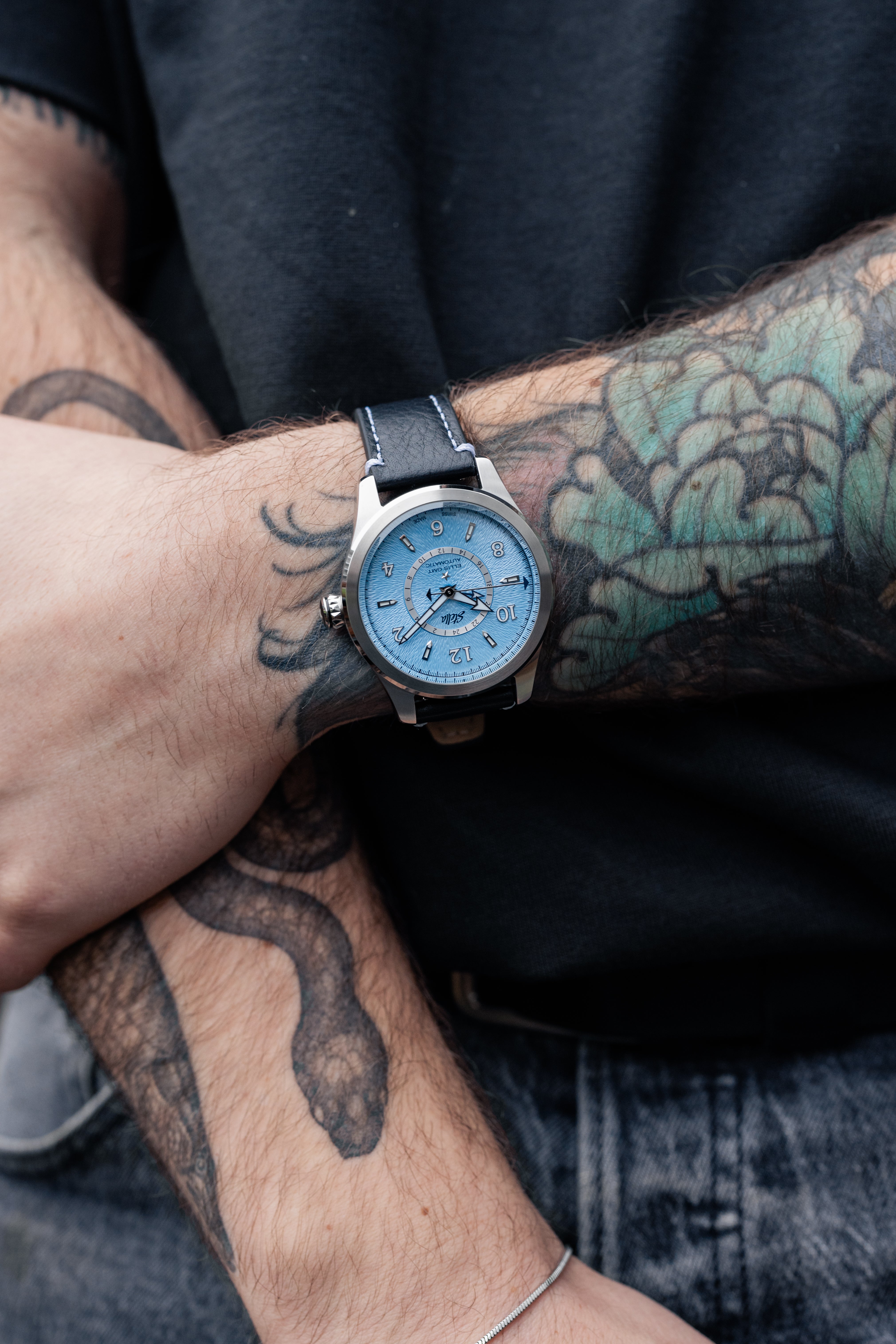 Person wearing a watch with a blue face on a tattooed arm.