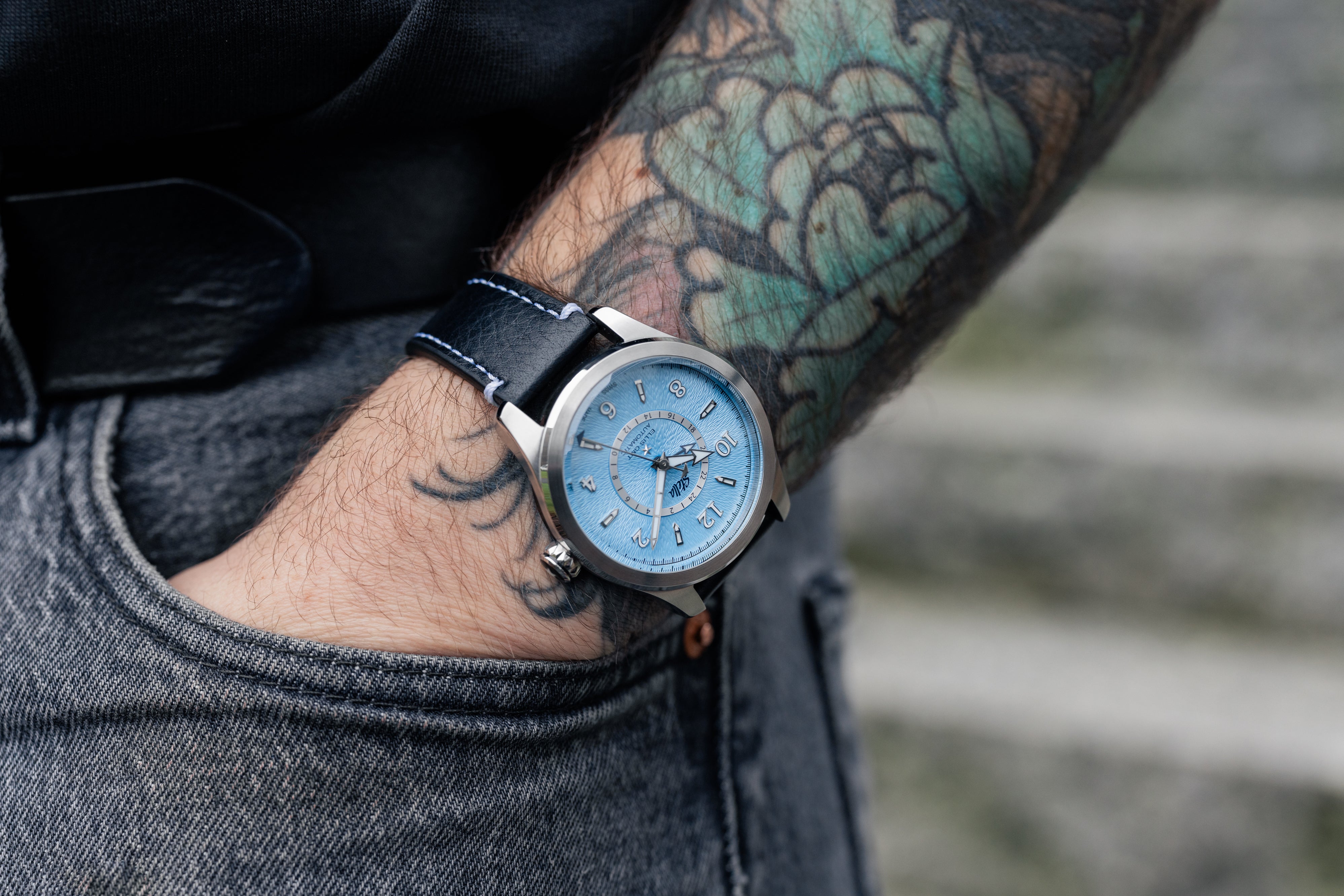 Person wearing a blue wristwatch with a tattooed arm on a blurred background