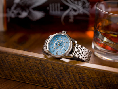 Silver watch with a blue face on a wooden surface next to a glass of whiskey.