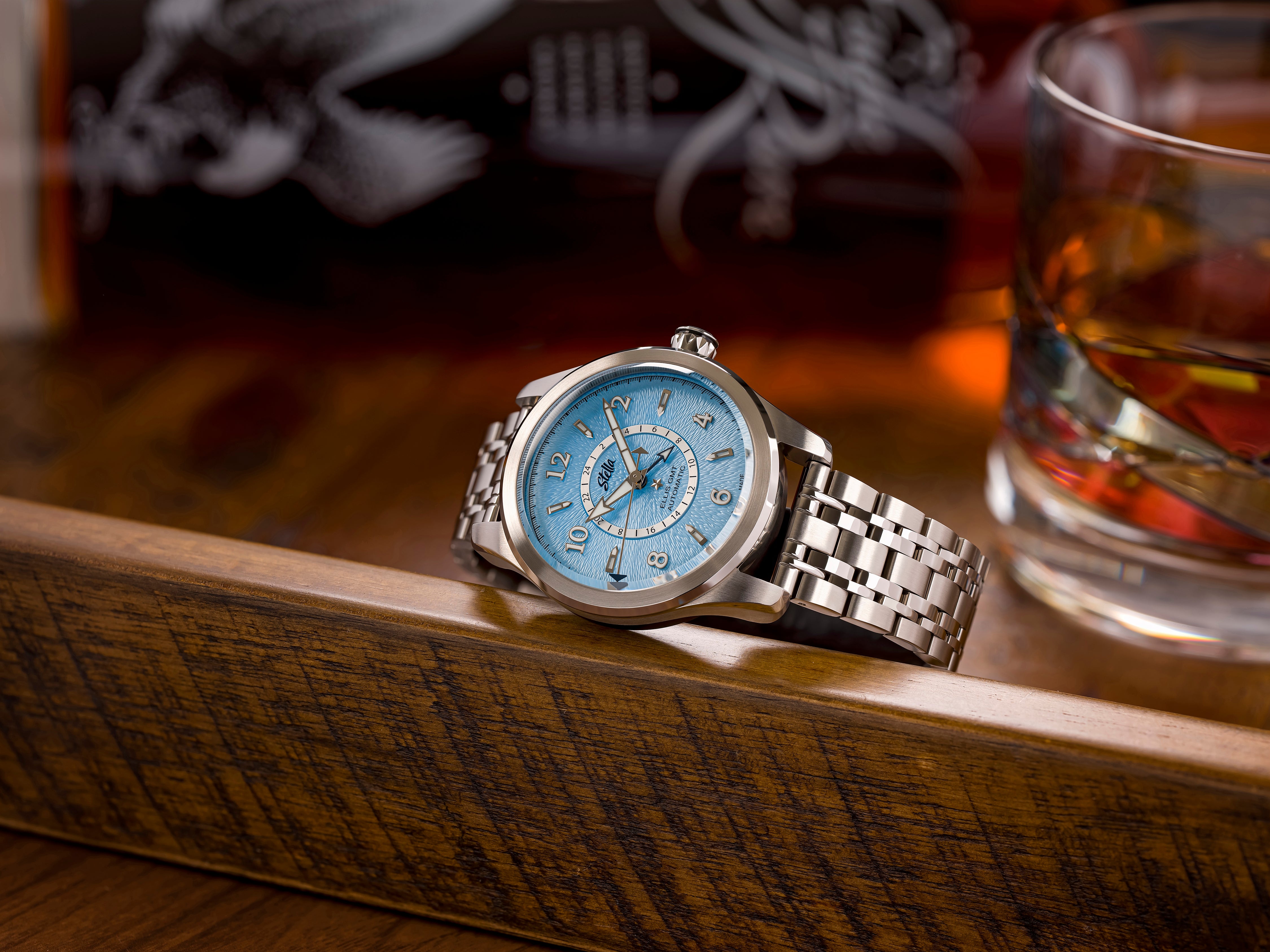 Silver watch with a blue face on a wooden surface next to a glass of whiskey.